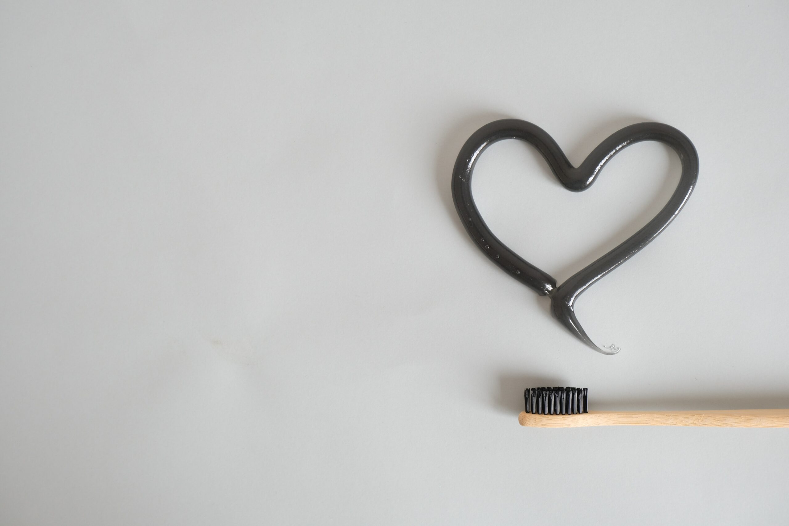 A wooden toothbrush is lying on a white countertop. Toothpaste is frosted in the shape of a heart next to it.